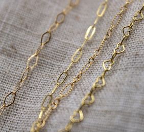Close-up of delicate gold chain necklaces with oval and heart-shaped links laid on textured beige linen