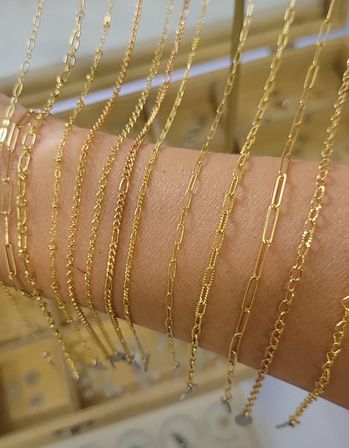 Close-up of shimmering gold chain necklaces in assorted delicate link styles draped across a forearm as a jewelry display, showcasing thin paperclip, rope and textured links