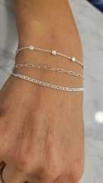 Close-up of a wrist wearing three stacked delicate silver chain bracelets — dotted bead chain, elongated link chain, and flat curb-style chain — photographed on a white marble surface.