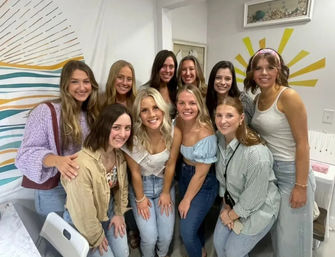 Smiling group of women posing indoors for a bachelorette-party photo, bride-to-be wearing a sash, casual jeans and tops, colorful wall mural and sunburst decor in the background.