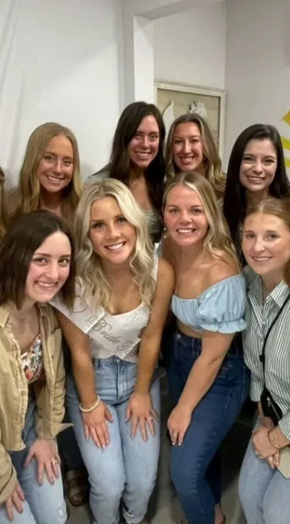 Smiling group of women posing indoors for a bachelorette-party photo, bride-to-be wearing a sash, casual jeans and tops, colorful wall mural and sunburst decor in the background.