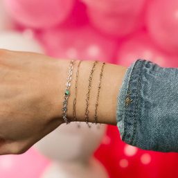 Close-up of a wrist wearing four delicate bracelets — three thin gold chains and a silver beaded bracelet with a small turquoise stone, denim sleeve and pink blurred background