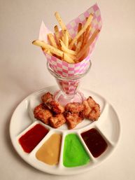 Crispy French fries in a pink checkered paper cone with golden fried chicken bites and a white platter of colorful dipping sauces