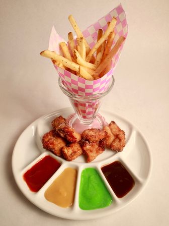 Crispy French fries in a pink checkered paper cone with golden fried chicken bites and a white platter of colorful dipping sauces