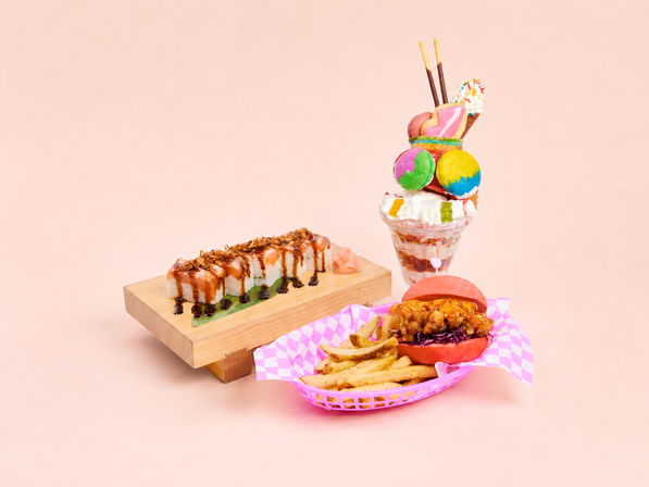 Colorful food trio on a pastel pink background: sushi rolls on a wooden board drizzled with sauce, a bright pink-bunned fried chicken slider with seasoned fries in a checkered basket, and an over-the-top milkshake stacked with macarons, cookies and whipped cream.