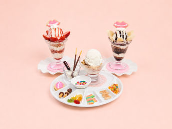 Three ice cream parfaits and a central vanilla scoop with assorted toppings—fresh strawberries, banana slices, chocolate sauce, colorful sprinkles, gummy candies and playful lip-shaped cookies—on a pastel pink background.