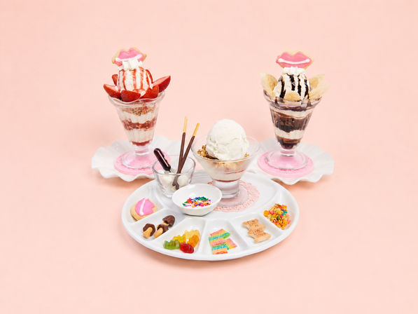 Three ice cream parfaits and a central vanilla scoop with assorted toppings—fresh strawberries, banana slices, chocolate sauce, colorful sprinkles, gummy candies and playful lip-shaped cookies—on a pastel pink background.