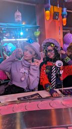 Two people in colorful kawaii-punk outfits posing at a neon-lit indoor party or café booth — one in a lavender sweater and hat with star necklace, the other in red-and-black layered accessories; purple balloons and hanging lanterns in background.