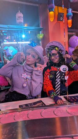 Two people in colorful kawaii-punk outfits posing at a neon-lit indoor party or café booth — one in a lavender sweater and hat with star necklace, the other in red-and-black layered accessories; purple balloons and hanging lanterns in background.