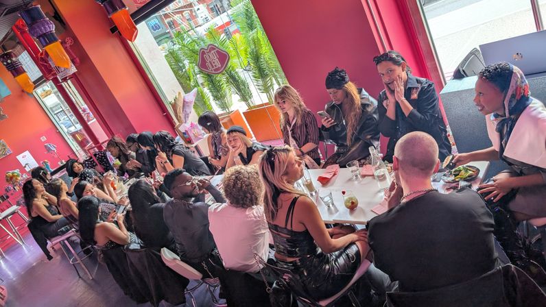 Diverse group dining at a lively pink cafe brunch, seated at a long table by large windows with tropical plants and colorful pendant lights in an urban city setting.