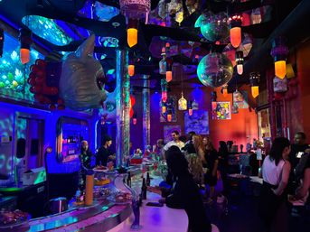 Vibrant neon-lit conveyor-belt sushi bar with colorful hanging lanterns and disco balls, oversized whimsical face sculptures and reflective columns, patrons gathered at the counter under blue and purple mood lighting.