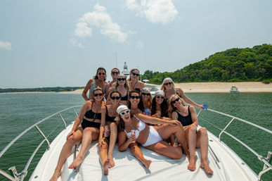 Group of friends in swimsuits and sunglasses laughing and holding drinks while lounging on the bow of a white yacht near a sandy beach and green coastal hillside under a sunny summer sky