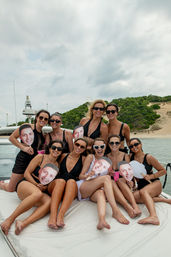 Group of women in swimsuits posing on a yacht near sandy coastal dunes, holding cutout face masks for a fun summer boat party