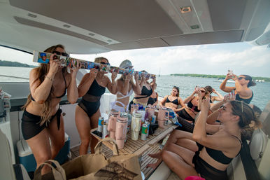 Group of women in swimsuits enjoying a summer yacht party on a coastal bay, taking photos and using a decorated shot-ski over a table of drinks and tumblers