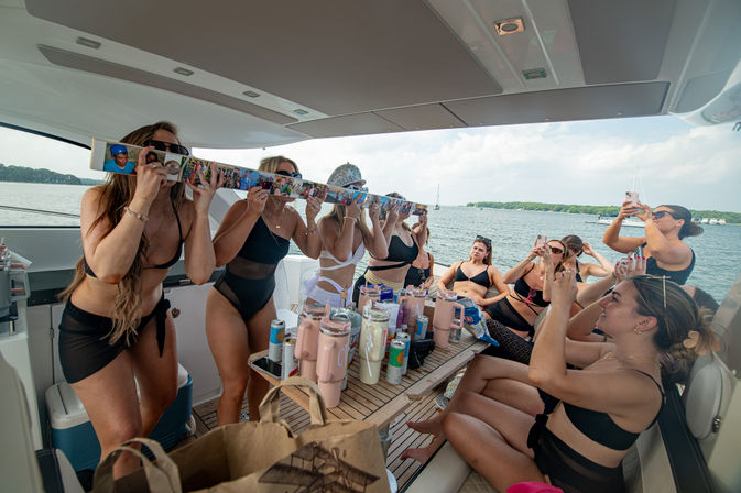 Group of women in swimsuits enjoying a summer yacht party on a coastal bay, taking photos and using a decorated shot-ski over a table of drinks and tumblers