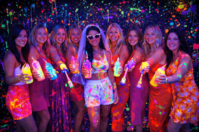 Smiling group of women at a blacklight neon paint party, bride wearing veil and heart-shaped sunglasses holding fluorescent paint bottles in front of a splattered glow backdrop
