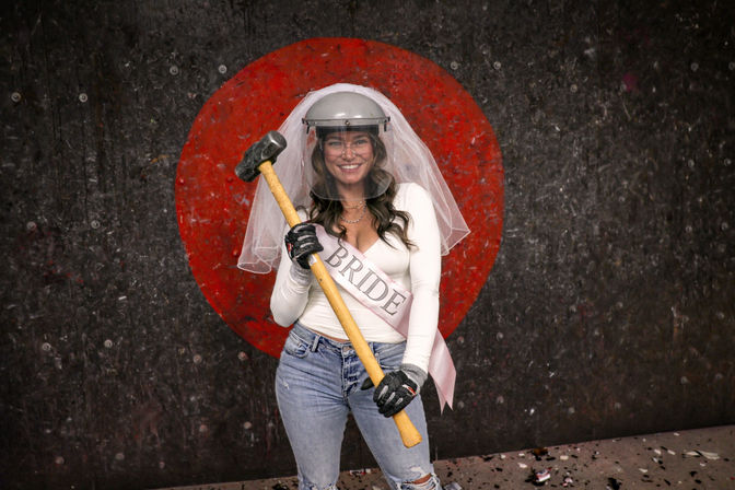 Bride-to-be in veil and "BRIDE" sash smiling while holding a sledgehammer, wearing safety helmet and gloves in an indoor smash/rage room in front of a red target
