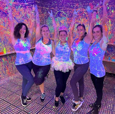 Group of five smiling women in neon tank tops and leggings pose with arms raised at an indoor UV blacklight paint party, covered in colorful fluorescent paint splatters.