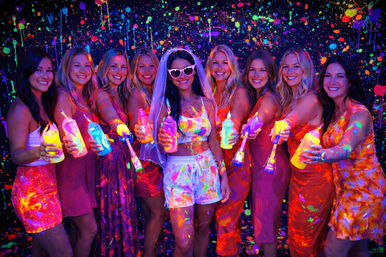 Smiling group of women at a blacklight neon paint party, bride wearing veil and heart-shaped sunglasses holding fluorescent paint bottles in front of a splattered glow backdrop