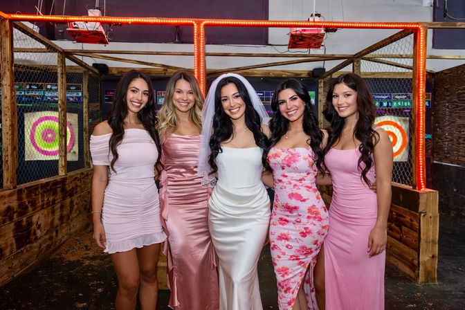 Bride in a white satin gown and veil smiling with four bridesmaids in pink dresses, posing inside a lit indoor axe‑throwing venue with wooden lanes and colorful target boards.