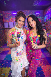 Two smiling women in a UV-lit splatter-paint studio, wearing a white lace dress and a pink dress, covered in neon paint and holding colorful paintbrushes.