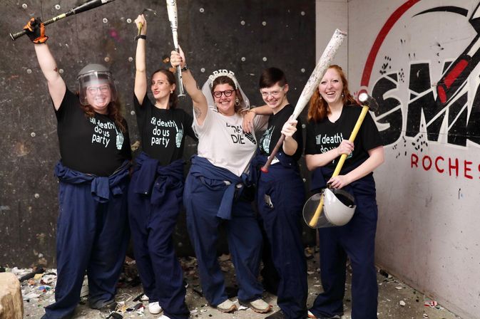 Group of five adults in a smash room wearing coveralls and face shields, posing with baseball bats and a sledgehammer; one person wears a bridal veil for a playful bachelorette celebration amid shattered debris.