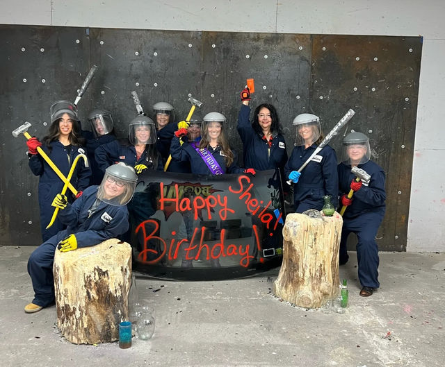 Group in coveralls and face shields posing with sledgehammers and crowbars in an indoor smash room for a birthday celebration, standing behind a black board painted "Happy Birthday" with wood stump props and scattered bottles.