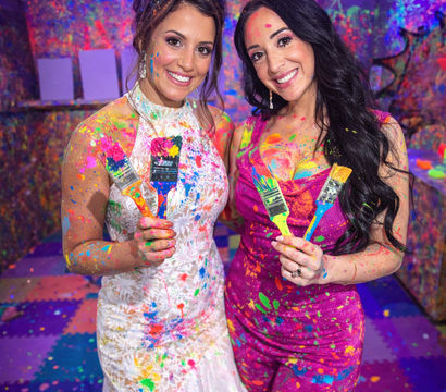 Two smiling women in a UV-lit splatter-paint studio, wearing a white lace dress and a pink dress, covered in neon paint and holding colorful paintbrushes.