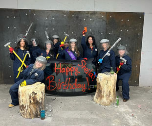 Group birthday smash-room scene: people in navy coveralls and face shields holding sledgehammers and crowbars behind a broken glass panel painted "Happy Birthday", with wood-stump props and scattered bottles.
