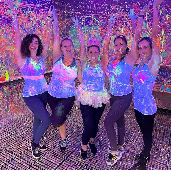 Group of five smiling women in neon tank tops and leggings pose with arms raised at an indoor UV blacklight paint party, covered in colorful fluorescent paint splatters.