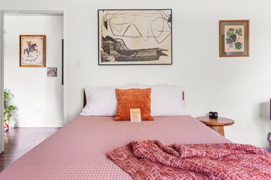 Cozy modern bedroom interior with a pink quilted bed, orange textured pillow and knit throw, abstract wall art above, wooden round nightstand and white walls.