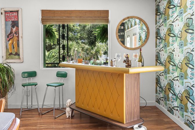 Sunlit retro home bar nook with mustard tufted counter, two green vinyl bar stools, mid-century patterned wallpaper, round mirror, wooden floor and leafy backyard view