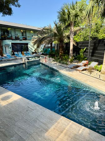 Sunlit resort-style courtyard with a deep-blue outdoor pool, bubbling fountain, palm trees, striped lounge chairs and blue deck chairs along a two-story building