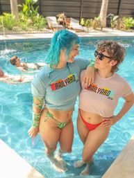 Two friends smiling and posing in the shallow end of a sunny backyard pool, wearing colorful bikini bottoms and cropped graphic tees — one with bright blue hair and the other in sunglasses, lounge chairs and tropical plants in the background.