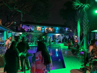 Vibrant nighttime poolside party in a tropical backyard venue with green and purple lighting, palm trees, guests mingling around an illuminated pool and a two-story balcony with live music.