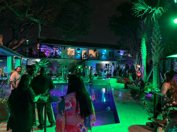 Vibrant nighttime poolside party in a tropical backyard venue with green and purple lighting, palm trees, guests mingling around an illuminated pool and a two-story balcony with live music.