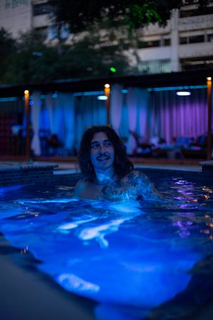 Person with long hair and chest tattoos relaxing and smiling in an urban outdoor pool glowing blue and purple under cabana and string lights at night
