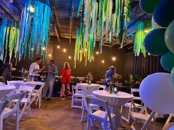 Festive indoor event space with industrial ceiling, blue and green hanging streamers, warm string lights and a balloon column; white-clothed round tables with floral centerpieces and guests mingling at a casual celebration.