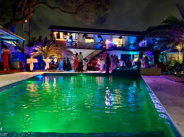 Nighttime poolside party in a tropical backyard venue with a glowing green pool, colorful LED lights, palm trees and guests mingling by a two-story house