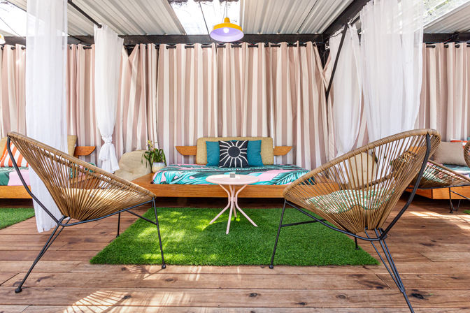 Sunlit covered patio lounge on a wooden deck with two woven rattan chairs, a low daybed with tropical-print cushions, striped curtains and a small round coffee table on artificial grass.