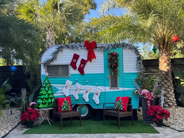 Teal-and-white vintage camper decked out for Christmas in a palm-lined tropical backyard with garlands, wreath and red bow, monogram stockings, two wicker chairs with festive pillows, poinsettias and a pink flamingo