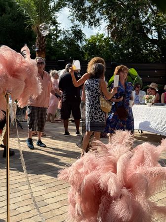 Sunlit outdoor garden party on a brick patio with palm trees, guests mingling by a lace-covered table, pink feather centerpieces and floating bubbles.