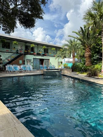 Tropical courtyard with turquoise outdoor pool, palm trees, striped lounge chairs and a two-story pastel-green motel balcony under a bright blue sky.