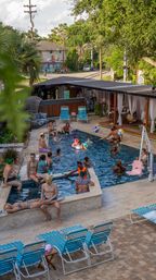 Outdoor urban pool party in a hotel-style courtyard — adults playing with colorful beach balls, lounging on blue-striped chairs and under cabanas amid palm trees and greenery.