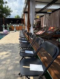 Sunny poolside patio set for an outdoor event: long row of black folding chairs with programs and wooden fans along the deck beside a pool, covered cabana with white curtains, wicker lounge chairs with striped pillows and a bar in the background.