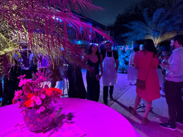 Vibrant outdoor tropical-themed nighttime party with neon pink and blue lighting, close-up of a cocktail table with a floral centerpiece and palm fronds, guests mingling in the background