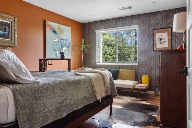 Sunlit modern-rustic bedroom with orange accent wall and patterned gray wallpaper, gray quilted bed, wooden mid-century bench under a window overlooking green trees, cowhide rug and wooden dresser.