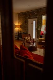 Cozy urban loft living room with exposed brick wall, warm lamp glow, wooden floors, woven leather lounge chair with striped pillow, round leather ottoman, red sofa cushions and a colorful curtain by a glass-panel door