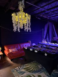 Warehouse-style event lounge with a hanging white floral chandelier glowing warm yellow, red leather sofa, buffet trays on a black-clothed table, purple uplighting and string lights