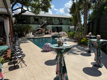 Sunny outdoor poolside courtyard set for a tropical-themed party with palm-print high-top tables and floral centerpieces, folding lounge chairs along the deck, wicker daybed and inflatable ball by a blue pool, two-story building balcony and palm trees.